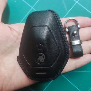 May include: Black leather car key fob cover with a matching key ring. The fob cover has a textured surface and a car logo. The key ring has a silver metal ring and a leather strap.