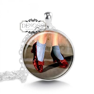 May include: A silver-toned circular pendant necklace featuring a graphic print of legs wearing light blue socks and sparkling red shoes with bows. The text DENOSS JEWELRY & GIFT is printed in black on a white rectangular background above the image.
