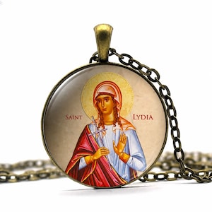 May include: A bronze-toned pendant necklace featuring a round charm with an image of Saint Lydia. The image depicts a woman in a white and blue robe, holding a cross. The words "SAINT LYDIA" are printed on the charm.