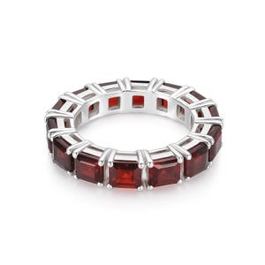 May include: A silver ring with a continuous band of rectangular, deep red gemstones. The gemstones are set closely together, creating a full circle of color. The ring's metal setting is bright and polished.