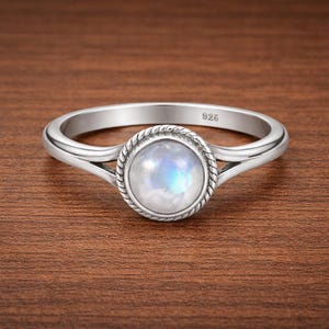 May include: A silver ring featuring a round moonstone with iridescent qualities. The stone is encircled by a braided border, and the ring has a split shank design. The band is marked "925". The ring is displayed on a brown wooden surface.