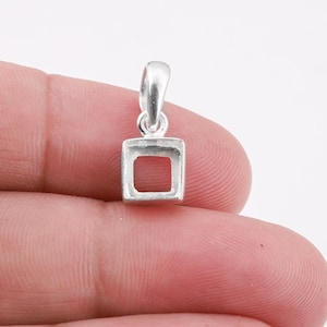 Peut inclure: Un petit pendentif argenté avec un design carré à cadre ouvert. Le pendentif a une boucle en haut pour une chaîne. Le cadre carré est vide, créant une esthétique minimaliste. Le pendentif est tenu par les doigts d'une personne.