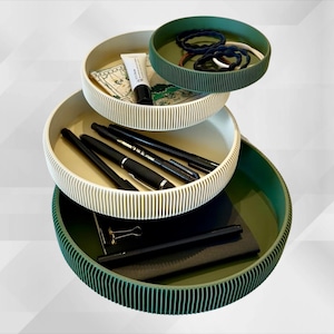 Könnte beinhalten: Ein mehrstöckiger Organizer mit drei runden Tabletts in Creme und Grün. Die Tabletts enthalten Stifte, ein Notizbuch, Schönheitsprodukte und Haargummis. Die Tabletts haben ein geripptes Außendesign.