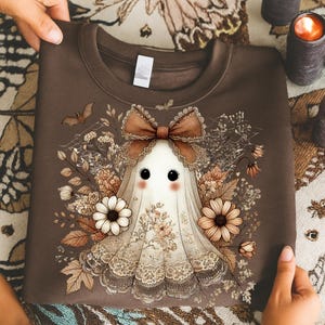May include: A brown sweatshirt featuring a whimsical ghost design. The ghost is adorned with a brown bow, floral accents, and a lace-trimmed veil. The design includes a bat and various flowers in shades of brown, beige, and white.