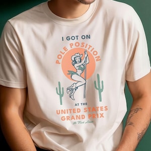 May include: A cream-colored short-sleeve t-shirt featuring a graphic of a person in a cowboy hat on a pole, with the text I GOT ON POLE POSITION THE UNITED STATES GRAND PRIX, surrounded by two green cacti and a large orange circle.