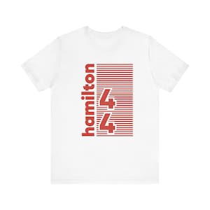 May include: White t-shirt with a red graphic. The graphic features the word "hamilton" in a vertical orientation, along with the number "44" in a rectangular design with horizontal lines. The shirt has short sleeves and a crew neck.
