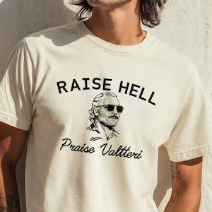 May include: Cream-colored t-shirt with the text "RAISE HELL" above a black and white illustration of a man wearing sunglasses. Below the illustration, the text reads "Praise Valtteri".