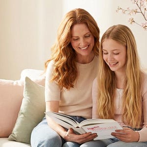 Pu&ograve; includere: Una donna e una ragazza sono sedute su un divano chiaro, che leggono un libro insieme. La donna ha lunghi capelli rossi e indossa un top chiaro e jeans blu. La ragazza ha lunghi capelli biondi e indossa un maglione rosa. Sul divano ci sono cuscini rosa e verdi.