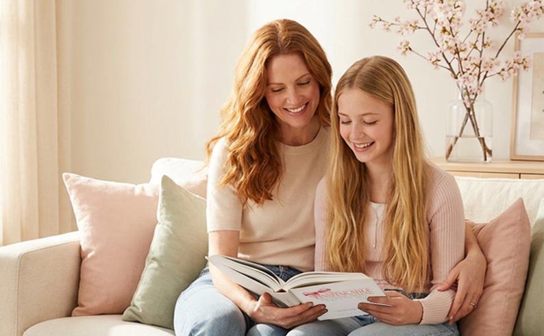 Pu&ograve; includere: Una donna e una ragazza sono sedute su un divano chiaro, che leggono un libro insieme. La donna ha lunghi capelli rossi e indossa un top chiaro e jeans blu. La ragazza ha lunghi capelli biondi e indossa un maglione rosa. Sul divano ci sono cuscini rosa e verdi.