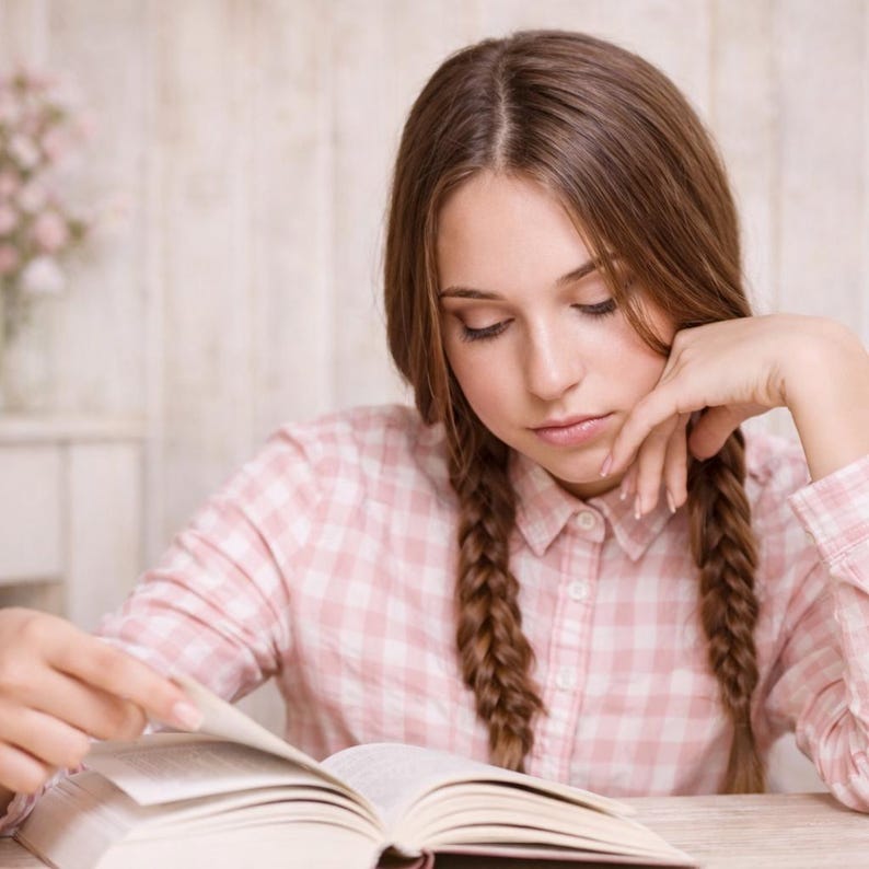 Pu&ograve; includere: Una persona con lunghe trecce marroni che indossa una camicia a quadri rosa e bianca, legge un libro. La persona &egrave; concentrata sulle pagine aperte, con la mano sul mento.