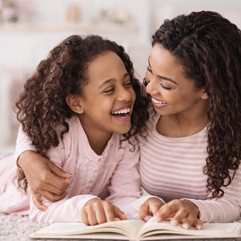 Pu&ograve; includere: Una madre e una bambina ridono mentre leggono un libro insieme. La bambina indossa una camicia rosa chiaro a maniche lunghe, e la madre indossa una camicia a righe rosa e bianca a maniche lunghe. Il libro &egrave; aperto e lo guardano entrambe.