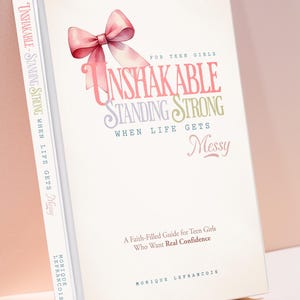 Op de afbeelding: Een boek getiteld "Unshakable Standing Strong When Life Gets Messy" met een roze strik illustratie. De cover bevat tekst in verschillende lettertypen en kleuren, waaronder "Unshakable", "Standing Strong" en "Messy". Het boek is een geloofsgerichte gids voor tienermeisjes.
