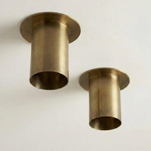May include: Two brass-colored cylindrical light fixtures with a circular base. The fixtures are mounted on a white ceiling. The cylindrical portion of each fixture is approximately 4 inches in height.