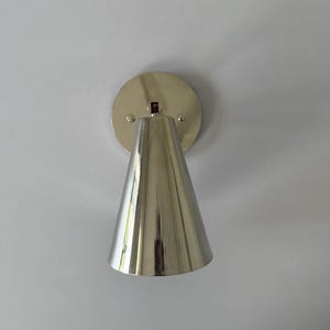 Op de afbeelding: Een gepolijste zilveren wandlamp met een conische kap en een cirkelvormige achterplaat. De lamp is aan een witte muur bevestigd, reflecteert licht en voegt een moderne touch toe aan het interieur.