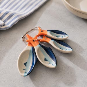 May include: A set of four ceramic measuring spoons, each designed as a fish. The spoons feature white, blue, and orange colours and are linked by a metal chain. The spoons are displayed on a grey surface, with a blue and white striped cloth visible.