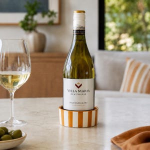 May include: A bottle of Villa Maria Sauvignon Blanc wine from New Zealand sits in a round, white and orange striped ceramic coaster. Beside it is a clear glass filled with white wine and a small bowl containing several green olives.
