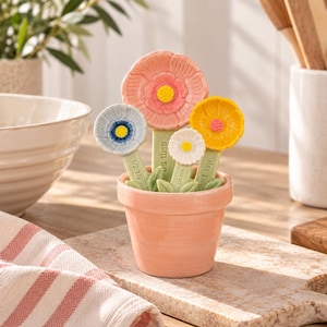 May include: A set of four flower-shaped measuring spoons in a small peach-coloured ceramic pot. The spoons feature a large pink flower, a yellow flower, a blue flower, and a white flower, each with green stems marked with measurements in tablespoons and teaspoons.