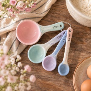 May include: A set of four ceramic measuring spoons connected by a metal ball chain. The spoons are pastel-coloured with pink, mint green, lavender, and light blue bowls. They are labelled 1 TBSP, 1 TSP, 1/2 TSP, and 1/4 TSP in black text.