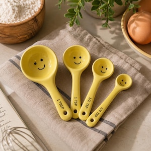 May include: A set of four yellow ceramic measuring spoons with black smiley faces printed in the bowls. The handles are labelled 1 TBSP, 1 TSP, 1/2 TSP, and 1/4 TSP in black text. Each spoon features a small circular hole at the end of the handle.