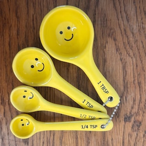 May include: A set of four yellow ceramic measuring spoons with black smiley faces. Each spoon is labeled with a measurement: 1 TBSP, 1 TSP, 1/2 TSP, and 1/4 TSP. The spoons are attached with a metal chain.