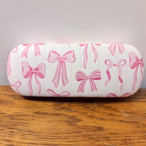 May include: An oval-shaped glasses case with a white background and a pattern of pink bows. The bows are in various styles, set against vertical pink stripes. The case is on a wooden surface.