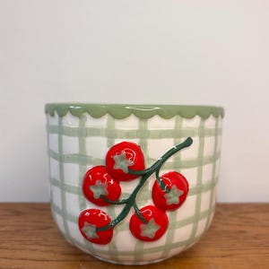 May include: A ceramic bowl with a green and white checked pattern and a scalloped rim. The bowl features a branch of red tomatoes with green star-shaped accents. The bowl is on a wooden surface.