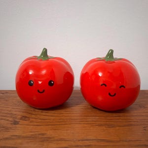 May include: Two red ceramic tomato-shaped ornaments with green stems and smiling faces. The glossy tomatoes sit on a wooden surface against a white background. These decorative items add a touch of whimsy to any kitchen.