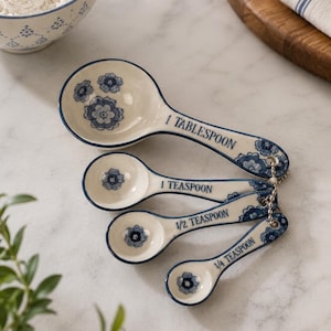 May include: A set of four ceramic measuring spoons with a blue and white floral pattern. Each spoon is labelled with its measurement: 1 Tablespoon, 1 Teaspoon, 1/2 Teaspoon, and 1/4 Teaspoon. The spoons are linked by a metal chain.