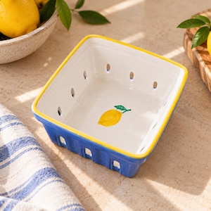 May include: A square ceramic berry basket with a bright blue exterior and a white interior. The rim is painted yellow, and a single yellow lemon with a green leaf is painted on the bottom. The sides feature small, evenly spaced ventilation holes.