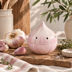May include: A ceramic garlic keeper shaped like a bulb of garlic with a white and pink streaked finish. The container features a pointed top, a two-piece design, and small ventilation holes around the middle to allow for airflow.