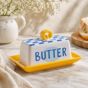 May include: A rectangular ceramic butter dish with a bright yellow base and a white lid featuring a blue checkered pattern on top. The word BUTTER is printed in blue letters on a light blue oval background. A yellow butter curl sits on the lid.