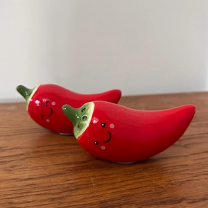 May include: A pair of red ceramic chilli pepper-shaped salt and pepper shakers with green stems and smiling faces. They sit on a wooden surface and have small white dots for cheeks.