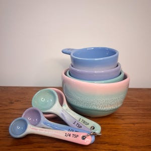 May include: A set of pastel-coloured ceramic measuring cups and measuring spoons. The set includes a stack of four bowls in shades of blue, purple, and green. Three measuring spoons are visible, labelled 1 TBSP, 1 TSP, 1/2 TSP, and 1/4 TSP.