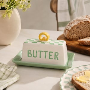 May include: A rectangular ceramic butter dish with a white base and a light green chequered pattern on the lid. The word BUTTER is printed in green on the front. A small yellow loop handle sits on top of the lid, which rests on a light green tray.