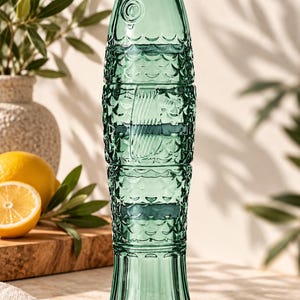 May include: A tall, translucent emerald green glass pitcher shaped like a fish. The exterior features a textured scale pattern across the body, a defined fin detail on the side, and a flared base. The neck is narrow with a simple circular rim.