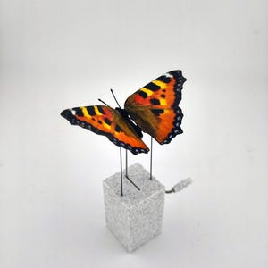 Könnte beinhalten: Eine detaillierte Skulptur eines Schmetterlings mit orangefarbenen, schwarzen und gelben Flügeln. Der Schmetterling ist auf einem kleinen, strukturierten, grauen Würfel mit dünnen Drahtstützen befestigt. Die Flügel sind ausgebreitet.