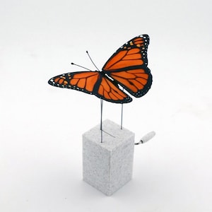 May include: A Monarch butterfly sculpture with orange and black wings, perched on a small, gray, cube-shaped base. The butterfly is supported by thin metal rods. The background is plain white.