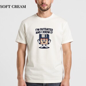 May include: Soft cream-colored t-shirt with the text "I'M OUTDATED AND I KNOW IT" above a cartoon floppy disk character. The floppy disk has a smiling face and small feet. The shirt is a short-sleeved crew neck.