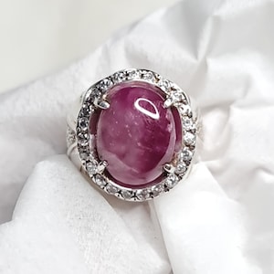 May include: A silver ring featuring a large, oval-shaped, deep red gemstone. The gemstone is surrounded by a halo of smaller, clear stones. The ring is set against a white background.
