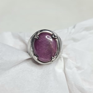 May include: A silver ring featuring a large, oval-shaped, purplish-red gemstone. The ring has a thick band and a bezel setting that securely holds the stone. The gemstone has a polished surface and a subtle, internal pattern.