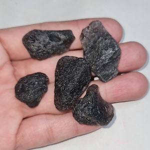 Puede incluir: Una colección de cinco piedras oscuras de forma irregular, posiblemente meteoritos, se sostienen en una mano. Las piedras varían en tamaño y tienen una superficie rugosa y texturizada. El color es negro carbón profundo con toques de gris.