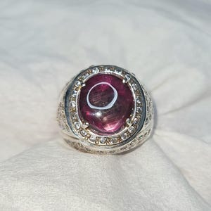 May include: A silver ring with a large, oval-shaped, deep pink gemstone at the center. The gemstone has a lighter pink circle in the middle. The ring is surrounded by small, clear stones.