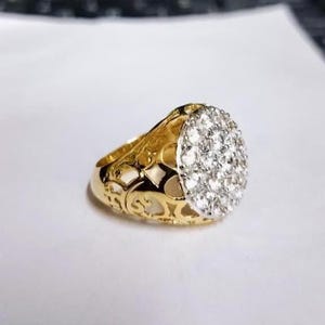 May include: A gold ring with a large, oval face set with numerous small, sparkling white stones. The band has an open filigree pattern. The ring is displayed against a white backdrop.
