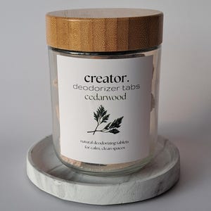 May include: A clear glass jar with a wooden lid, containing deodorizer tabs. The jar has a white label with the text "creator. deodorizer tabs cedarwood" and a sprig illustration. The jar sits on a round, marbled gray tray.