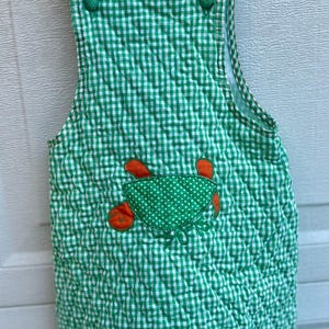 May include: Green and white gingham-patterned overall dress with a quilted texture. The dress features a green polka-dotted pocket with orange crab appliqués. It has green buttons on the straps.