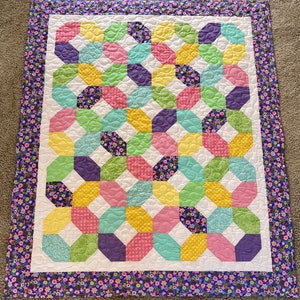 May include: A quilted baby blanket with a white background and a pattern of green, yellow, pink, and purple squares. The blanket has a purple and white floral border.