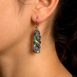 May include: A dangle earring featuring a rectangular, iridescent green and blue stone wrapped in dark copper wire. The wire forms intricate, swirling patterns around the stone, which hangs from a simple copper hook attached to the earlobe.