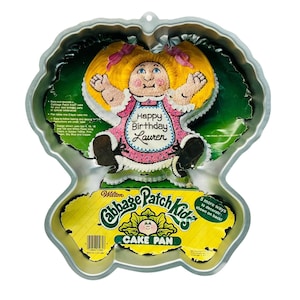 Könnte beinhalten: Eine Vintage Cabbage Patch Kids Kuchenform. Die Form hat die Form einer Cabbage Patch Kid-Puppe mit blonden Zöpfen und einem rosa Kleid. Die Worte "Happy Birthday Lauren" stehen auf der Schürze der Puppe. Die Form enthält den Text "Cabbage Patch Kids Cake Pan".