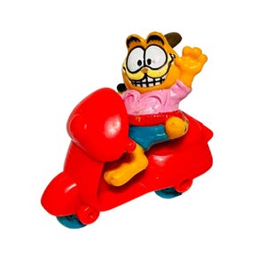 May include: A vintage Garfield figurine riding a bright red scooter. Garfield is orange with a pink shirt, blue shorts, and yellow hands and feet. The scooter has blue wheels and a yellow seat.