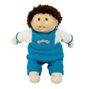 May include: A Cabbage Patch Kids doll with brown hair and brown eyes. The doll is wearing a blue knitted overall and a white and blue sweater. The Cabbage Patch Kids logo is on the front of the overall.